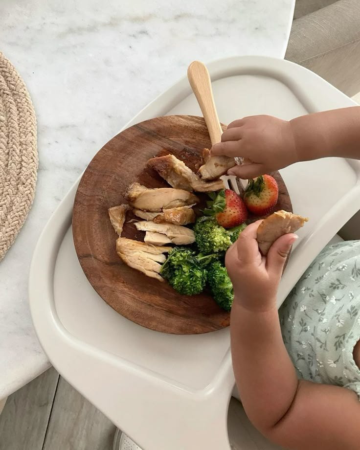 Criança pegando com as mãos frango, brócolis e morangos num prato de madeira