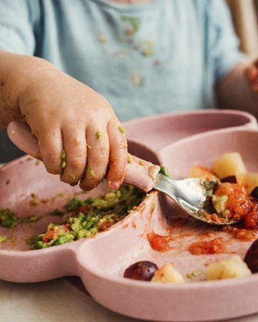 Criança comendo com colher num prato infantil rosa com legumes picados